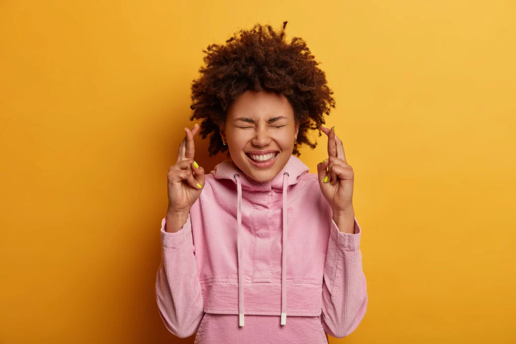 Uma pessoa com cabelos cacheados e um moletom rosa fica contra um fundo amarelo sólido, sorrindo com os olhos fechados e os dedos cruzados em ambas as mãos, transmitindo esperança ou expectativa.