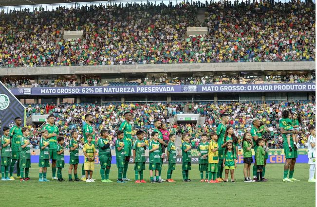 Times de futebol e crianças faziam fila em campo antes de uma partida do Brasileirão, com um estádio lotado de torcedores ao fundo.