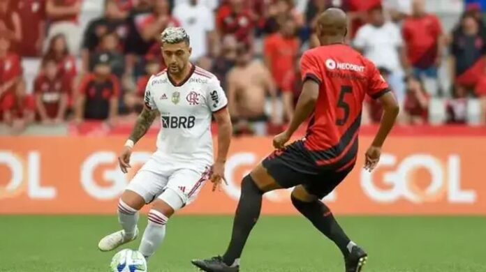 Dois jogadores de futebol do Flamengo e do Athletico Paranaense em uma partida, um driblando enquanto o outro se prepara para defender.