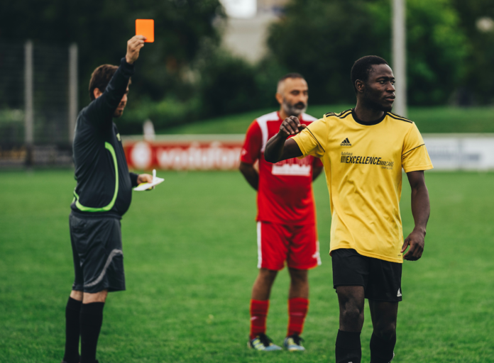 arbitro apontando cartão amarelo para jogador negro de camisa amarela.