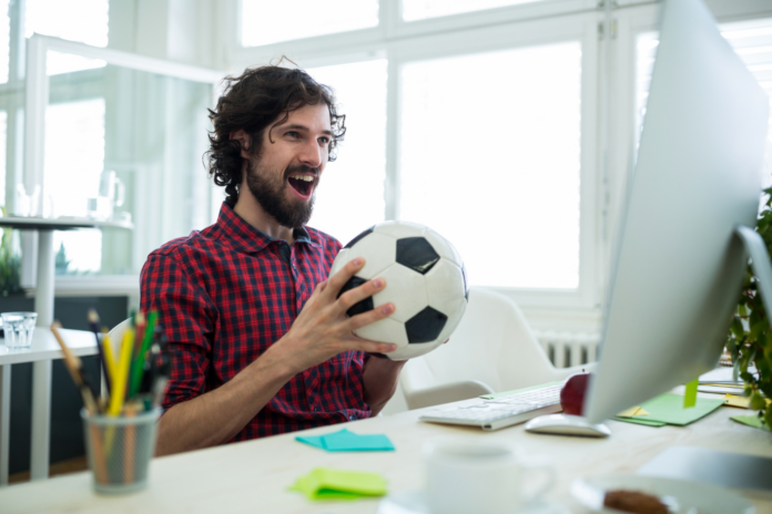 homem segura uma bola de futebol na mão enquanto vibra e sorri olhando para a tela do computador